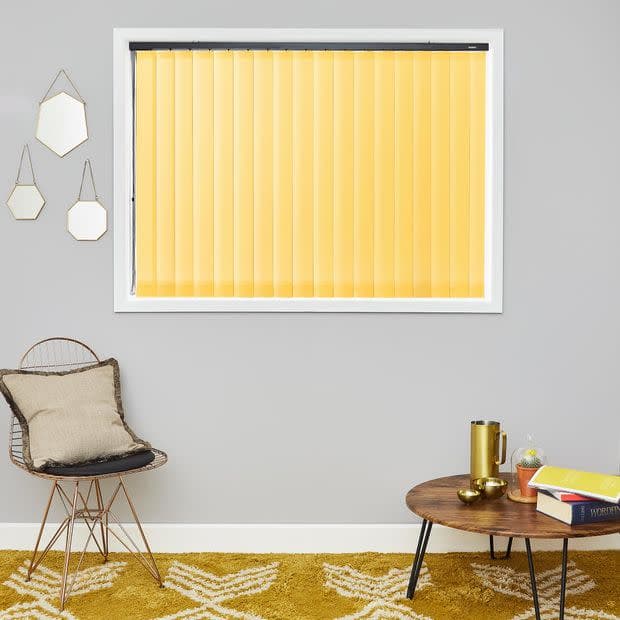 Yellow vertical blinds cover a centered rectangular window, closed and softly filtering daylight; set on a grey wall above a mustard rug, wire chair with cushion, round wooden coffee table.