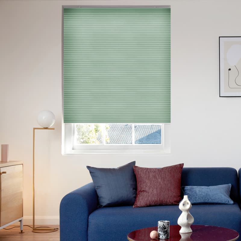 Mint-green pleated cellular blind, fully lowered with horizontal ribs, blocking most light; in a sunlit living room with a blue sofa, decorative pillows, floor lamp, sideboard, and partial rooftop view.