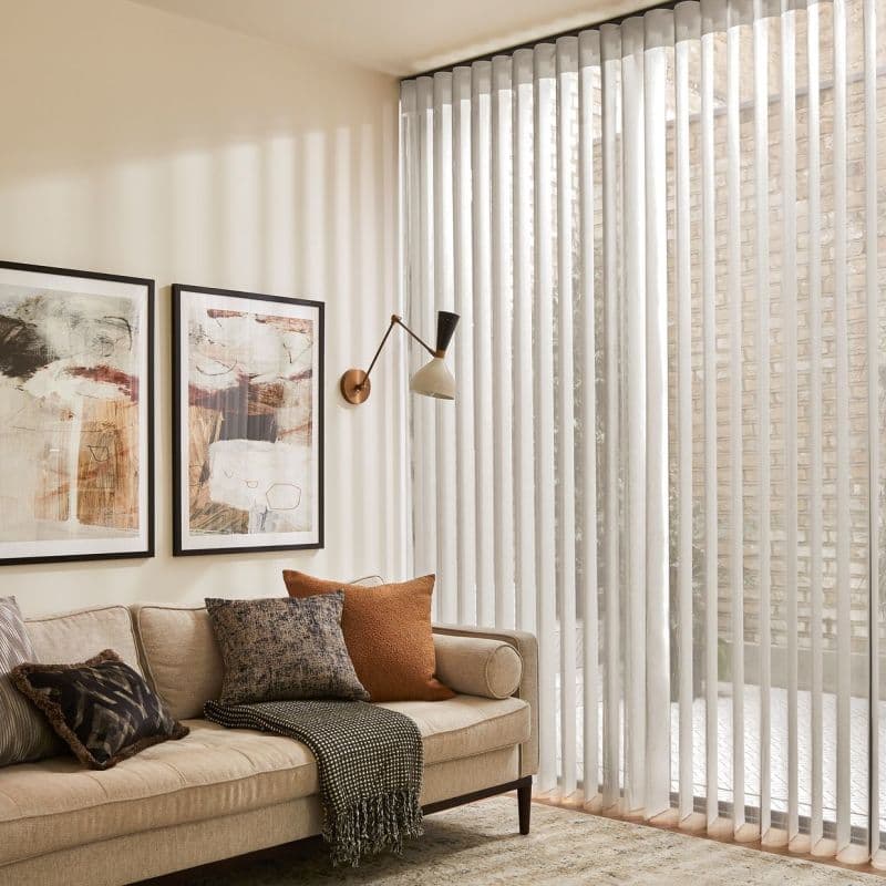 Sheer vertical blinds cover a floor-to-ceiling glass wall, partially open and filtering sunlight into vertical bands in a neutral living room with beige sofa, cushions and wall art.
