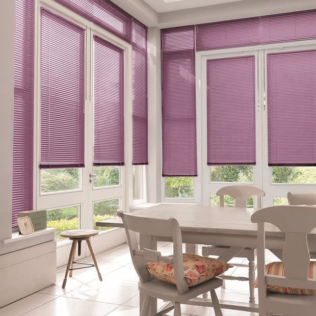 Purple horizontal venetian blinds covering tall bay windows and French doors, partially lowered and filtering soft daylight, in a bright white dining nook with table, chairs, cushions and garden view.