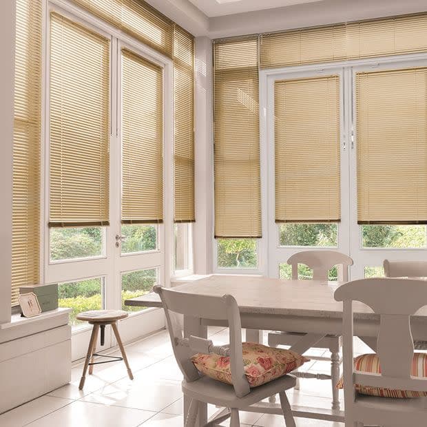Beige horizontal Venetian blinds covering tall patio windows and doors, slats mostly lowered; filtering soft daylight into a bright dining/sunroom with a wooden table, chairs, floral cushions, tiled floor.