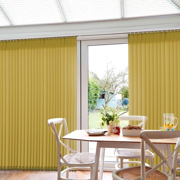 Yellow vertical blinds covering patio doors, mounted floor-to-ceiling with narrow slats, pulled aside to reveal central glass door. Dining table with chairs, flowers and breakfast items; bright daylight, garden view.