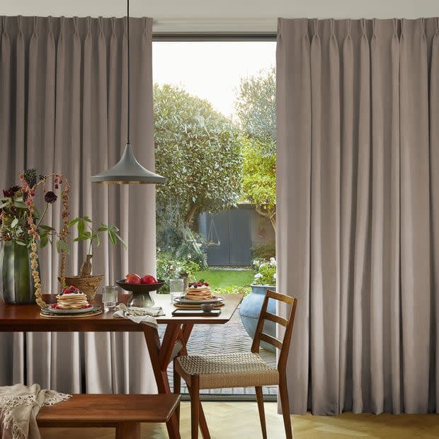 Floorâ€‘length pleated beige curtains, drawn to the sides and framing a glass door, letting daylight in; wooden dining table set for breakfast with pendant lamp, chairs, and backyard garden beyond.