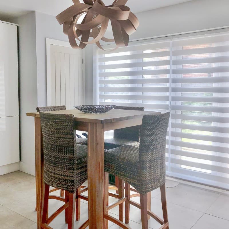 Dual-layer horizontal zebra roller blinds covering floor-to-ceiling sliding windows, alternating sheer and opaque bands, partially filtering bright daylight; dining nook with tall wooden table, woven bar chairs and sculptural pendant.
