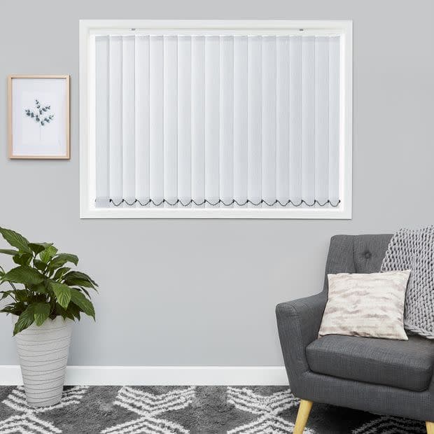 White vertical blinds cover the windowâ€”full-length slats with scalloped weighted bottomsâ€”closed, softly diffusing daylight. Context: minimalist gray living room with a gray armchair, potted plant, framed print, patterned rug.