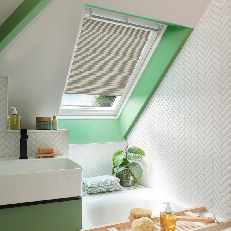 Pleated beige skylight blind, lowered halfway across a roof window, filtering daylight. Set in a green-framed sloped attic bathroom with white herringbone tiles, sink, tub, plant and bath accessories.