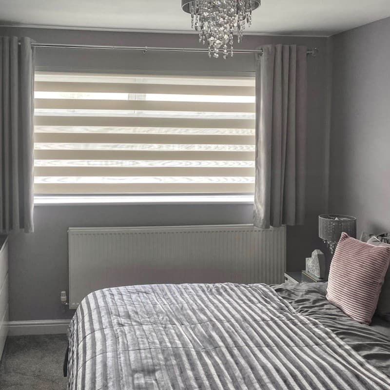 Zebra roller blind with alternating opaque and sheer horizontal bands lowered across a wide window, flanked by pleated grey curtains at the sides; filtering soft daylight into a grey-toned bedroom.