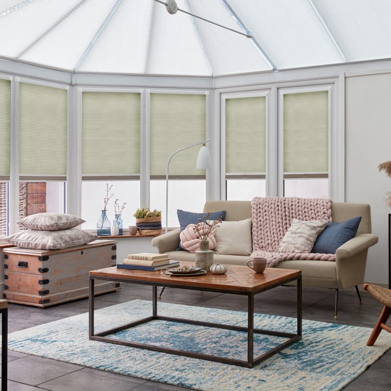 Pleated cellular blinds, pale green, partially lowered across tall white-framed conservatory windows, filtering daylight into a sunlit living area with beige sofa, wooden coffee table, and blue rug.
