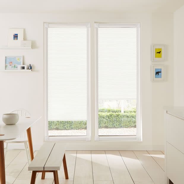 White pleated blinds covering two tall windows; the blinds are lowered, bottoms slightly raised to reveal garden hedge, filtering bright daylight into a minimalist dining space.