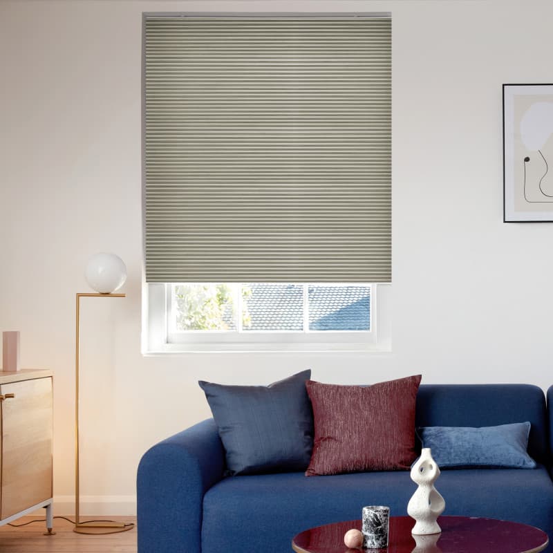 Beige narrowâ€‘pleated blind, lowered to cover most of the window with a bottom gap, filtering daylight. Bright living room with blue sofa, cushions, floor lamp and framed art; rooftop beyond.