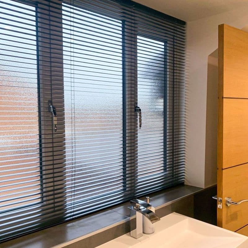 Dark horizontal Venetian blinds cover a three-panel frosted window to sill; slats slightly tilted, filtering daylight and obscuring view. Bathroom sink with chrome faucet in foreground beside wooden door.