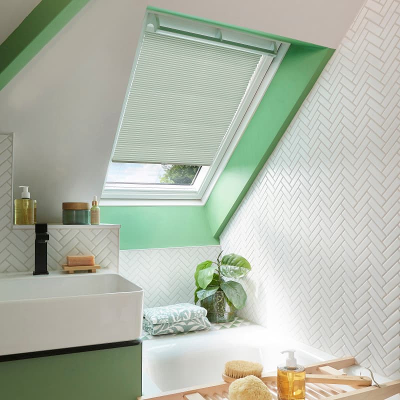 Pleated white cellular blind on an angled skylight, partially lowered and filtering daylight; sits above a green-framed attic bathroom with herringbone white tiles, bathtub, towel, potted plant, and toiletries.