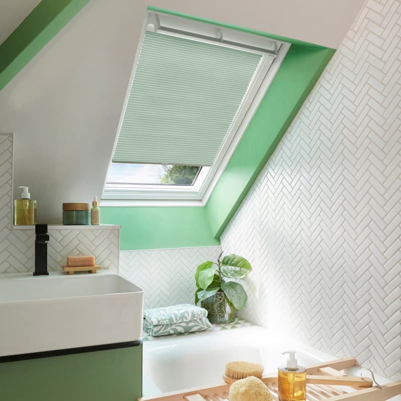 Mint-green pleated skylight blind, lowered and filtering daylight with a narrow open gap, in an angled bathroom window; white herringbone tiles, green trim, sink, tub, and potted plant nearby.