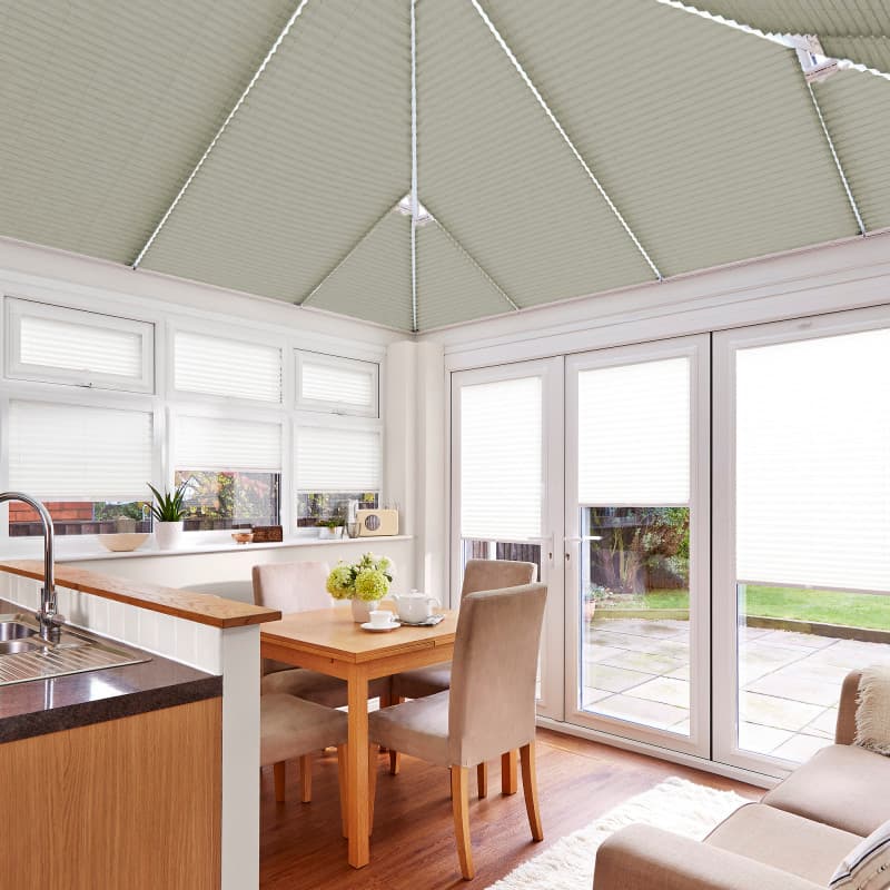 Pale pleated cellular blinds cover the conservatory roof, upper windows and French doors, honeycomb-textured and partially lowered, filtering bright daylight into a compact dining area overlooking a paved garden.