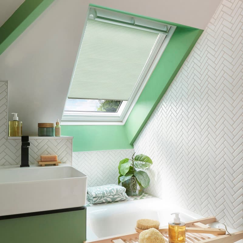 Pleated white skylight blind, partially lowered, filtering soft daylight; placed in a green-painted roof window recess above a bathtub in a bright attic bathroom with white herringbone tiles.