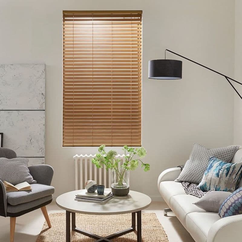 Wooden horizontal Venetian blinds, centered on the wall with narrow warm-toned slats, fully lowered and tilted to filter soft daylight; neutral living room with sofa, armchair, round coffee table, lamp.