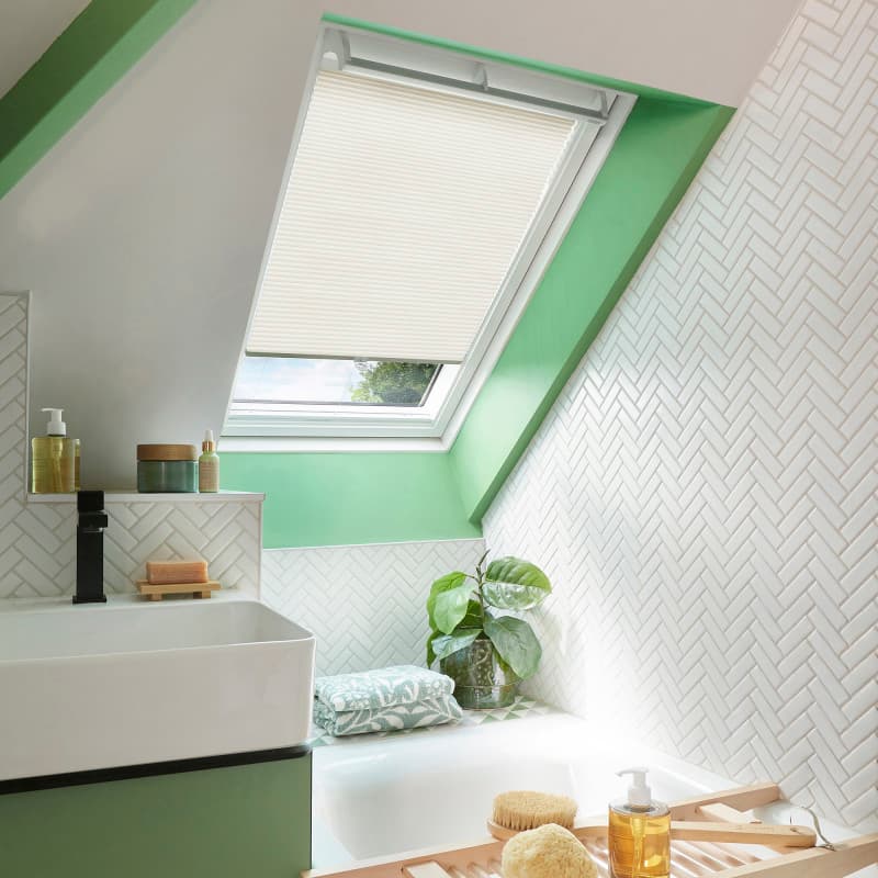 A beige pleated skylight blind, mounted inside an angled roof window, half-lowered, filtering daylight over a green-trimmed bathroom with white herringbone tiles, sink, bathtub, toiletries and a potted plant.