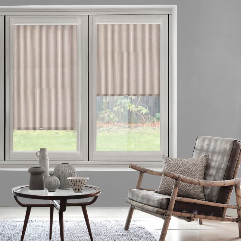 Striped beige pleated blinds cover the upper halves of twin windows, partially lowered and softly filtering daylight; modern living room with wooden-framed armchair, patterned cushion, small coffee table, lawn view.