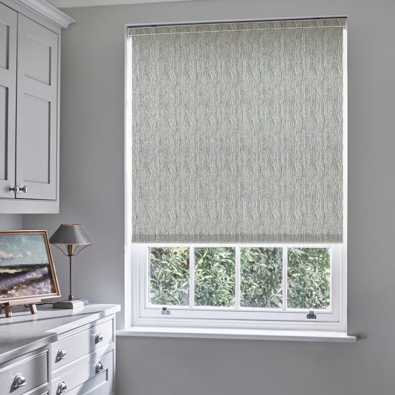 A patterned roller blind with subtle wavy grey lines, lowered to the sill, filtering daylight; in a grey room over a window above a dresser with lamp, garden visible.