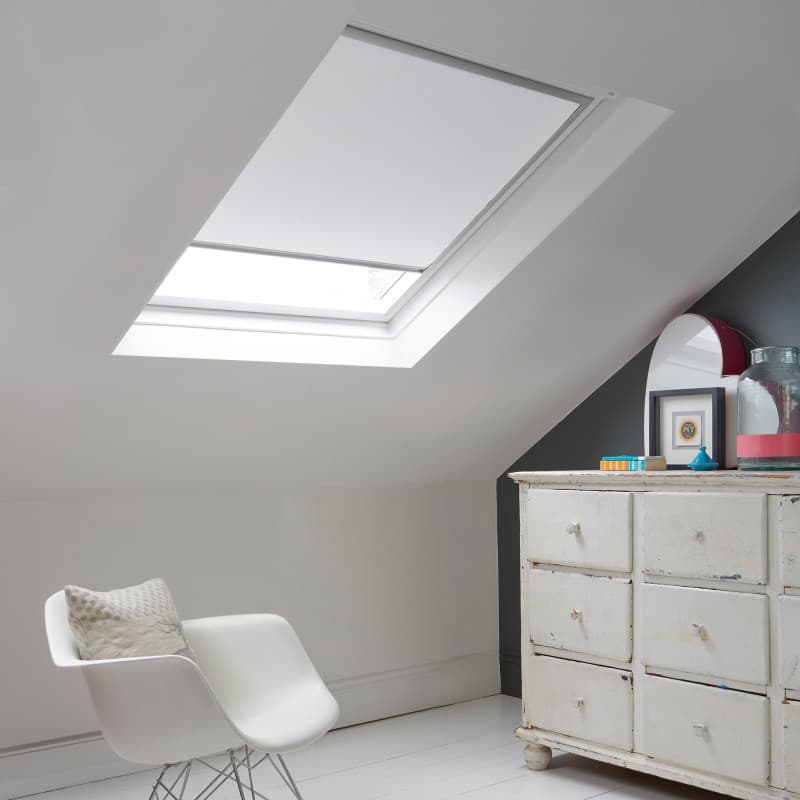 Room with white eaves wall, dark blue-grey wall to right with a shabby chic white drawer unit, a contemporary white chair, white painted floorboards and a skylight blind in white.