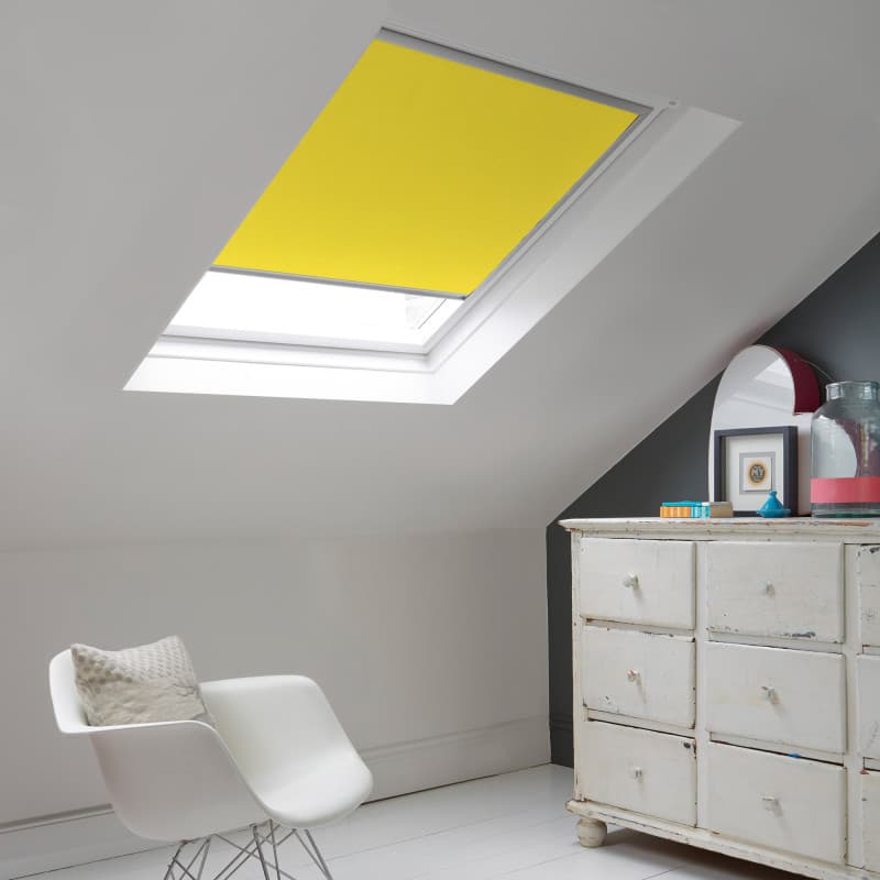 Room with white eaves wall, dark blue-grey wall to right with a shabby chic white drawer unit, a contemporary white chair, white painted floorboards and a skylight blind in sunshine yellow.