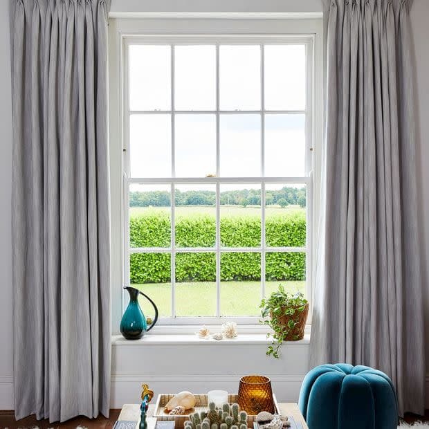 Floor-length pleated grey curtains frame a central multi-pane sash window; curtains are open, softly diffusing daylight into a cozy living room with a windowsill plant, jug, and hedged lawn view.