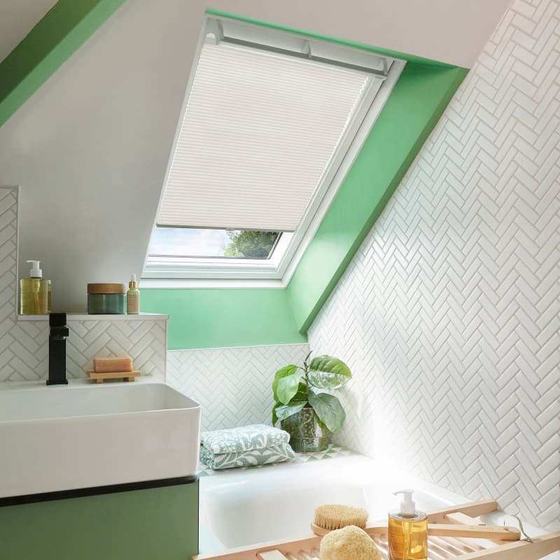 White pleated skylight blind, half-lowered and filtering soft daylight, covers a roof window above a sloped bathroom with mint-green trim, white herringbone tiles, sink, bathtub, plant and bath accessories.