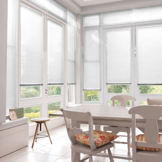 White horizontal venetian blinds cover tall bay windows, lowered and filtering soft daylight; overlooking a green garden, bright dining area with a white table, wooden chairs, and floral cushions.
