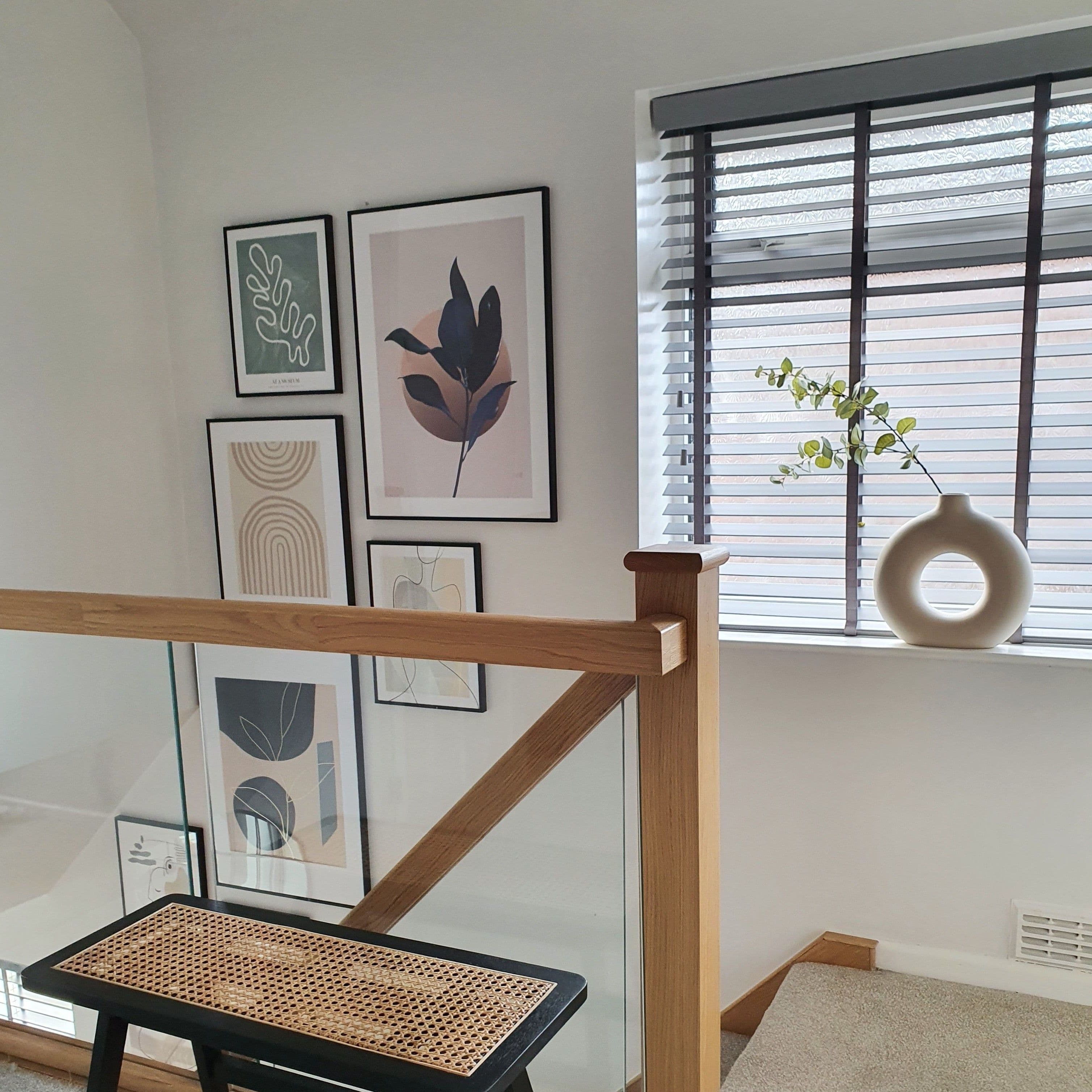 Horizontal slatted blinds on a landing window, partially tilted and filtering light; beside a wood-and-glass stair railing, framed abstract prints on the wall and a round vase on the sill.