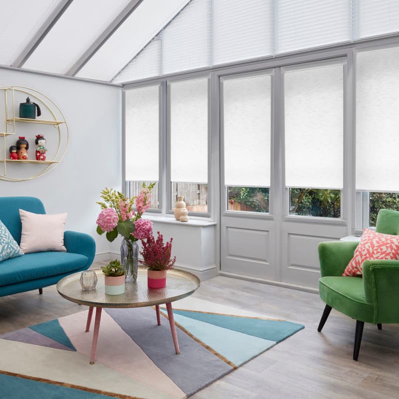 White roller blinds cover tall conservatory windows and triangular roof panels, lowered to diffuse daylight and obscure view; bright sunroom with teal sofa, green armchair, floral centerpiece and pastel rug.