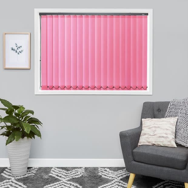 Pink vertical blinds covering a rectangular window, closed and slightly translucent, filtering daylight; set in a gray-walled living room with a gray armchair, patterned rug, potted plant, and framed art.