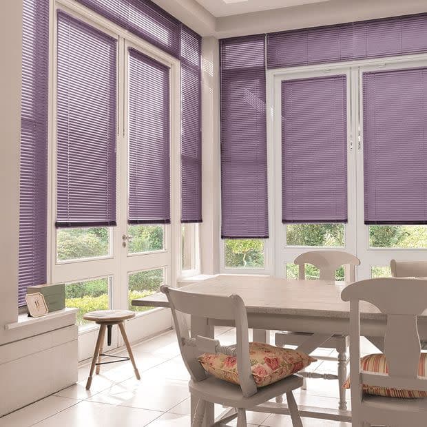 Purple horizontal Venetian blinds, lowered over tall French windows and doors with closed slats, filtering soft daylight into a bright tiled dining nook with white table, chairs, and garden views.