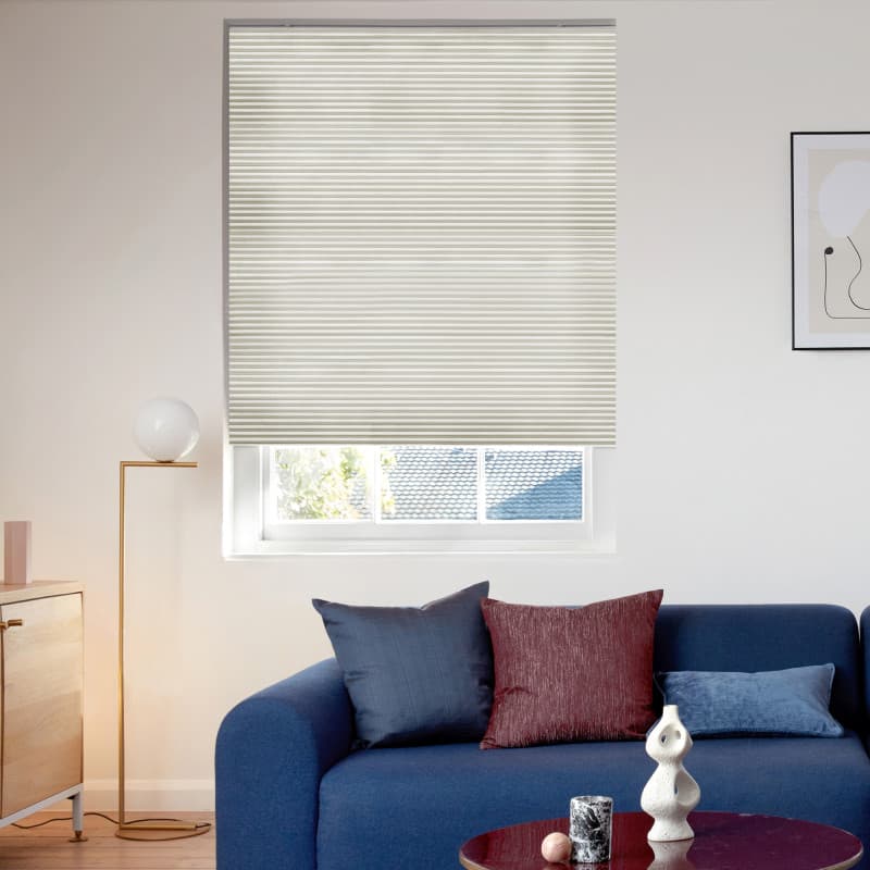 Cream pleated cellular blind lowered over window, filtering daylight and slightly raised to reveal lower panes; in a bright living room with a blue sofa, cushions, floor lamp, coffee table.