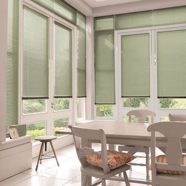 Green Venetian blinds on tall windows and patio doors, lowered and filtering daylight while leaving lower panes visible; bright sunroom dining area with white table, chairs, cushions, garden view.