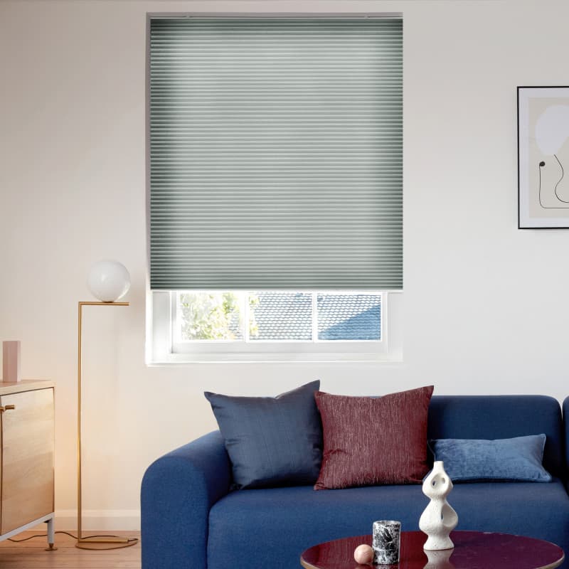 Pleated cellular blind, lowered almost fully with a narrow gap at the bottom and fine horizontal ribs, blocking and softly filtering daylight over a living room with blue sofa, lamp, and artwork.