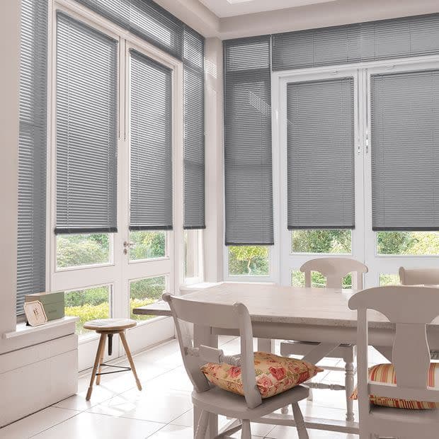 Grey horizontal Venetian blinds lowered over tall French doors and surrounding windows, filtering soft daylight into a bright dining nook with white wooden table, chairs and a visible garden view.