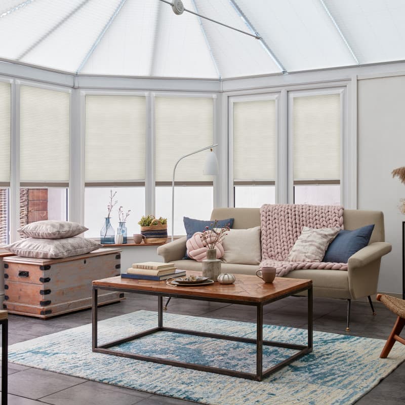 Cream honeycomb pleated blinds, lowered from the top leaving lower window panes exposed, diffusely filtering daylight across a bright conservatory-style living area with sofa, rug and coffee table.