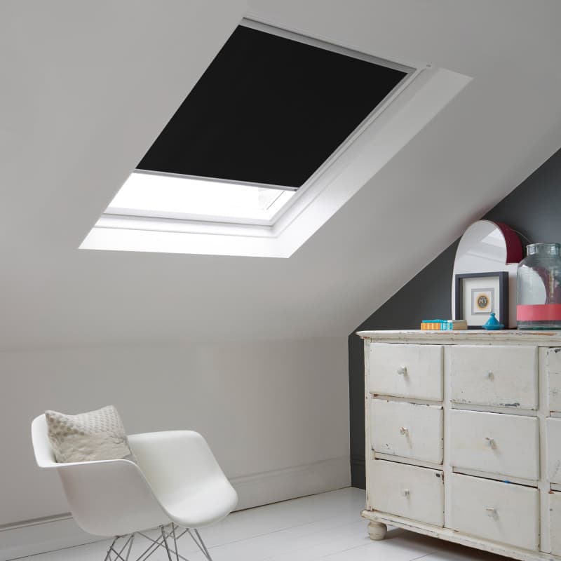 Room with white eaves wall, dark blue-grey wall to right with a shabby chic white drawer unit, a contemporary white chair, white painted floorboards and a skylight blind in black.