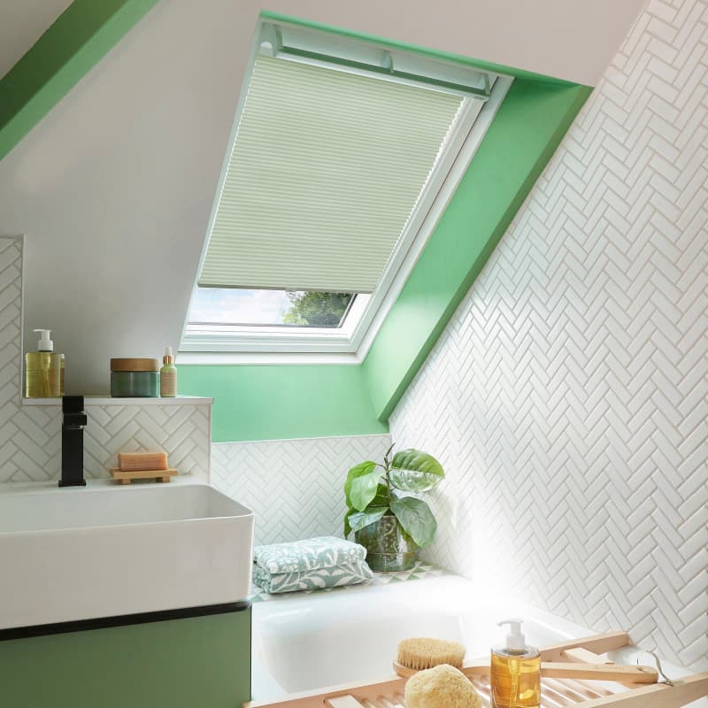 Pale cream pleated skylight blind, mounted in a sloped window and mostly lowered with a small gap, filtering daylight; bright bathroom with green trim, white herringbone tiles, sink and tub.