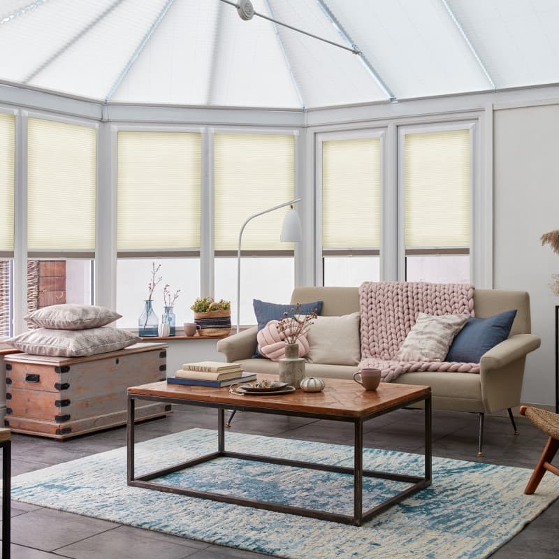 Cream honeycomb pleated blinds cover tall conservatory windows, lowered mid-height and softly diffusing daylight; cozy sunroom with beige sofa, pink knit throw, wooden coffee table and blue rug.