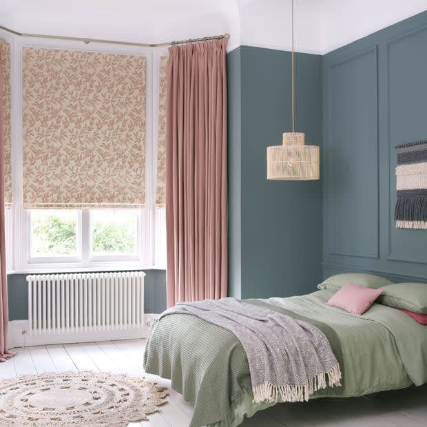 Floral-patterned roman blinds lowered over a bay window with long dusty-pink floor-length curtains drawn aside; they filter soft daylight into a teal-paneled bedroom with a green bed, woven pendant and round jute rug.
