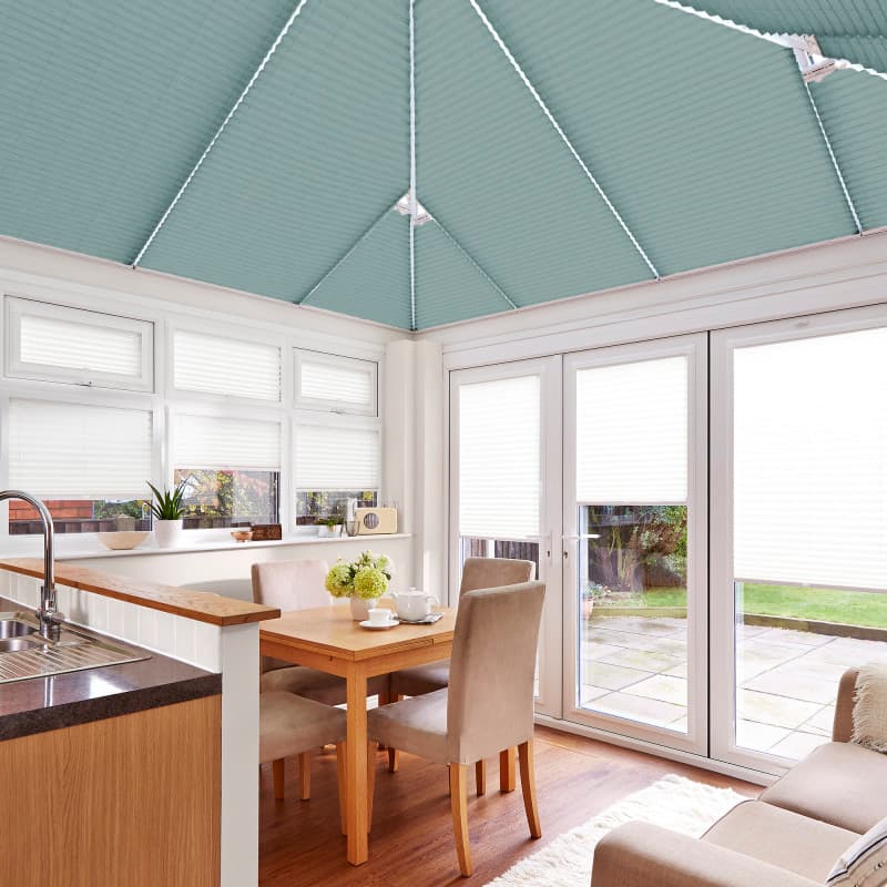 Teal pleated roof blinds cover the conservatory ceiling panels, meeting at the apex; fully lowered, diffusing light. White pleated window and door blinds closed; wood table, beige chairs, garden view.