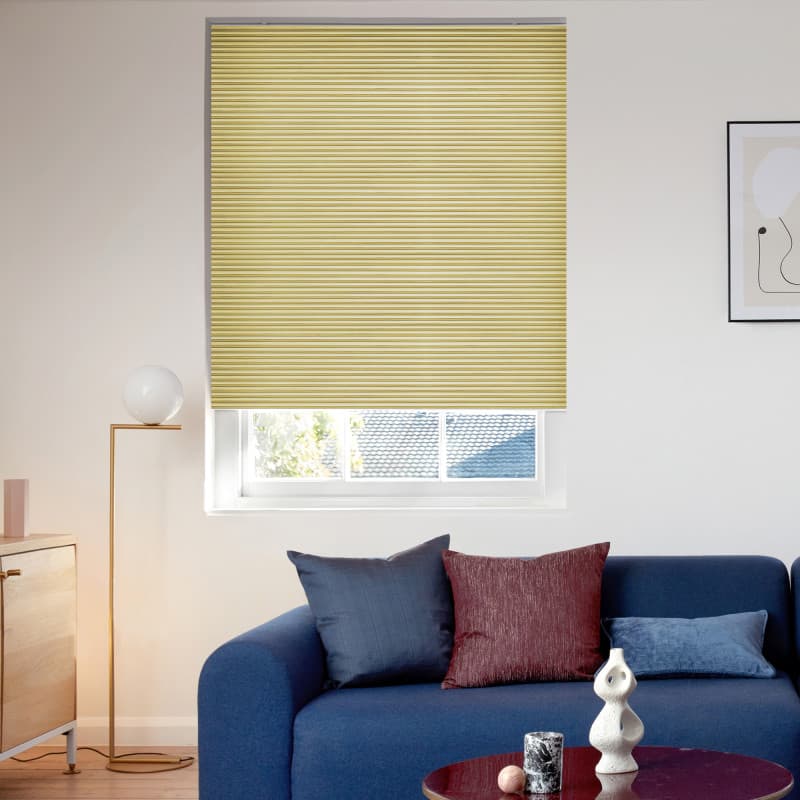 Yellow pleated cellular blind lowered over a window, closed and filtering daylight; small lower pane visible. Context: bright living room with navy sofa, patterned cushions, floor lamp and wall art.