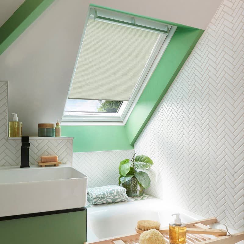 A cream pleated skylight blind, mostly lowered and filtering daylight through a slanted roof window; attic bathroom with bright green reveal, white herringbone tiles, sink, tub, plant and bath tray.