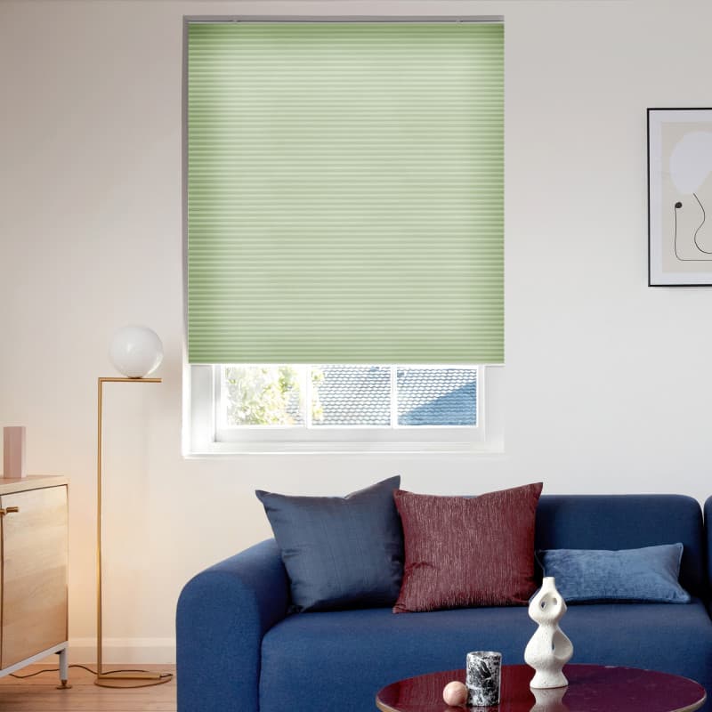 Light green cellular (honeycomb) blind, lowered almost fully with a narrow bottom gap, filtering soft daylight over a living-room window; blue sofa, colored cushions, floor lamp and framed art nearby.