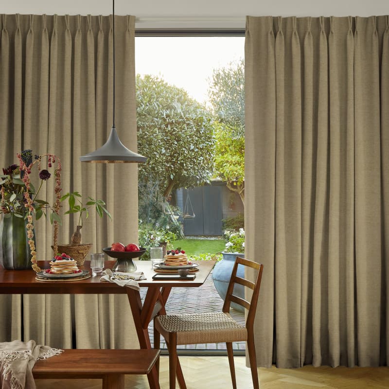 Beige pinch-pleat floor-length curtains framing a central opening, partially open and filtering daylight; in a dining room beside wooden table set for breakfast, pendant light and sunlit garden beyond.