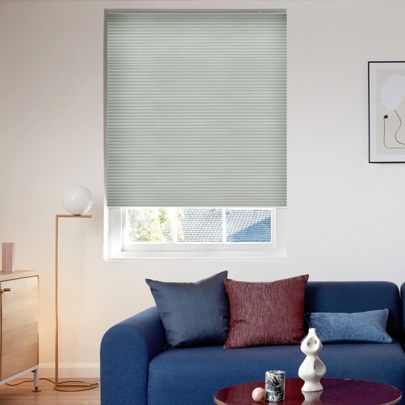 Light-green pleated cellular blind, lowered with horizontal ribs and a slightly raised bottom; filtering soft daylight into a living room with a blue sofa, cushions, and a brass floor lamp.