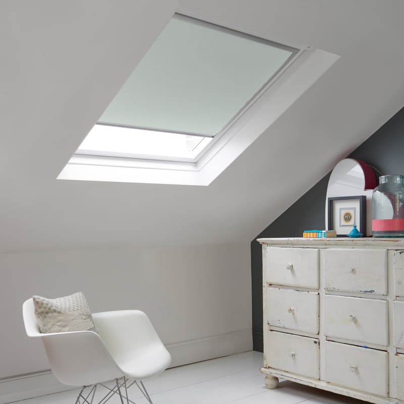 Room with white eaves wall, dark blue-grey wall to right with a shabby chic white drawer unit, a contemporary white chair, white painted floorboards and a skylight blind in pale grey.