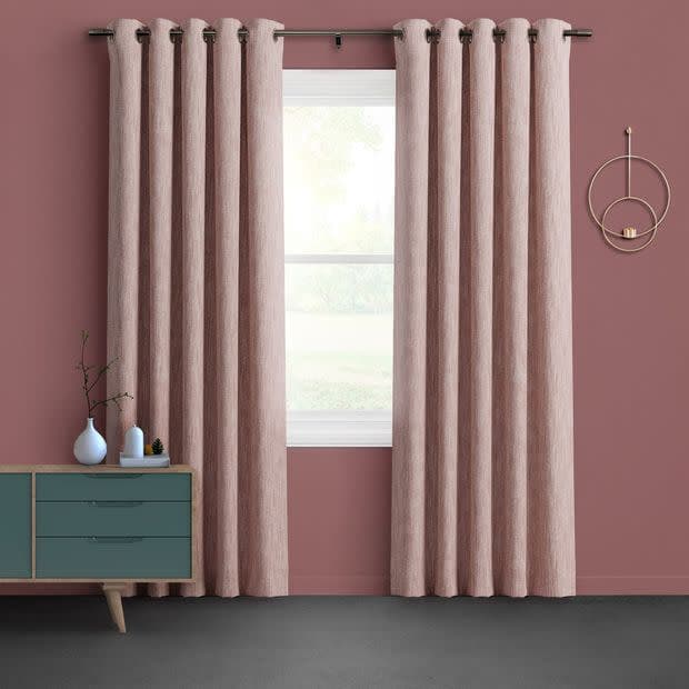 Dusty-pink grommet-top curtains hang flanking a central window, partially drawn and filtering soft daylight, set against a muted rose wall with a teal mid-century dresser and circular wall sconce.