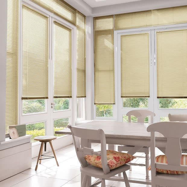 Beige horizontal venetian blinds covering tall corner windows, partly lowered and filtering soft daylight into a bright sunroom dining area with white table, chairs, floral cushions, and garden view.